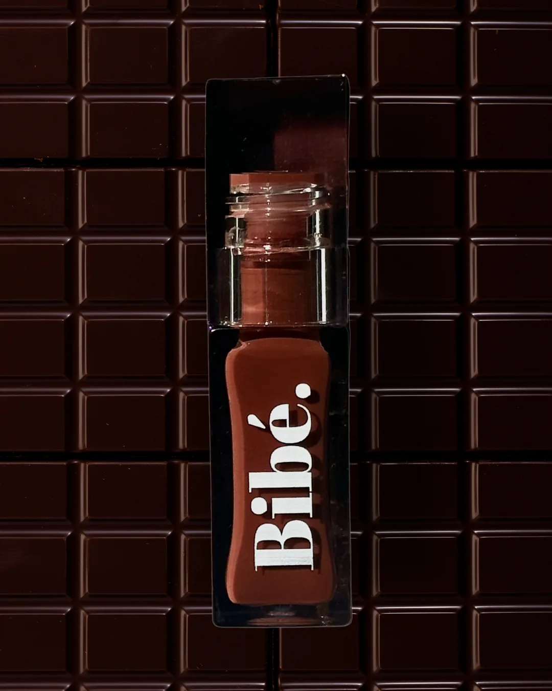 Bibé lip oil styled against a dark chocolate-textured backdrop