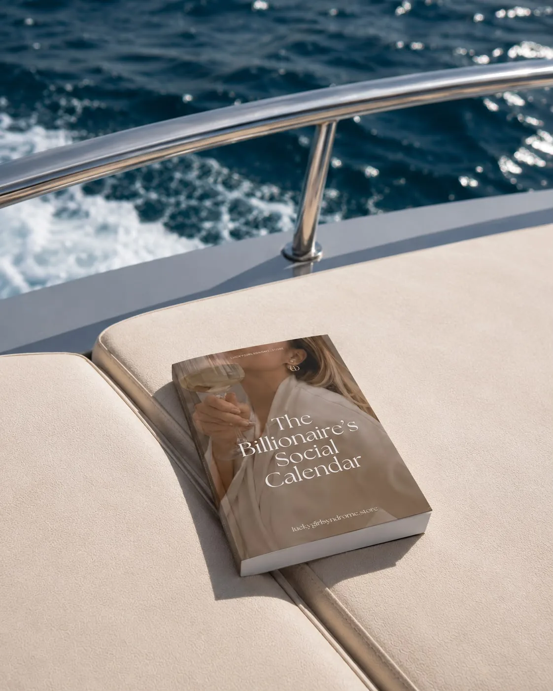 'The Billionaire's Social Calendar' paperback resting on a yacht cushion, ocean in the background